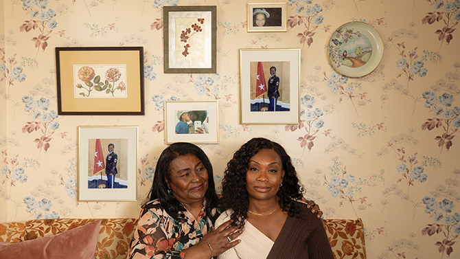 A Veteran and her mother sitting on a couch in front of floral wallpaper and a collage of portraits including some from her service