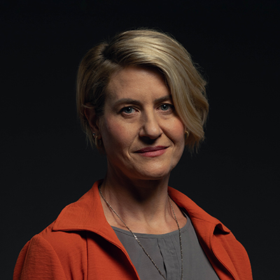 A middle aged blonde woman with a red jacket and looking directly at the camera with a subtle closed smile.